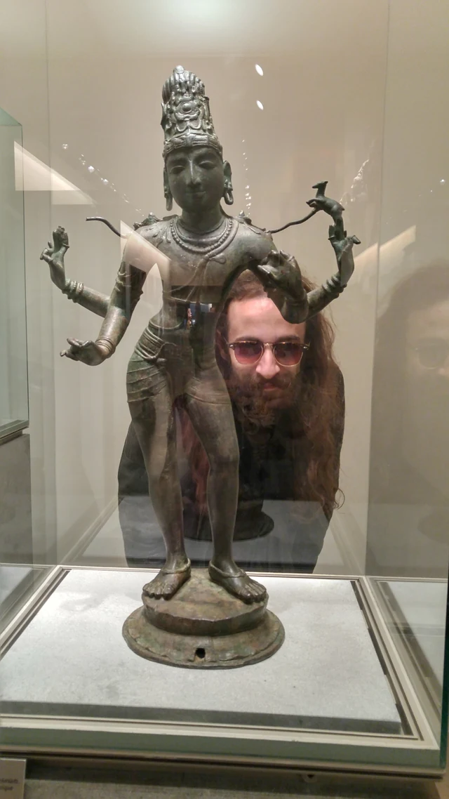A bearded person seen behind a small bronze dancing figure in a display case.