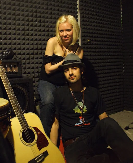 Two people posing together beside an acoustic guitar against soundproofing foam.
